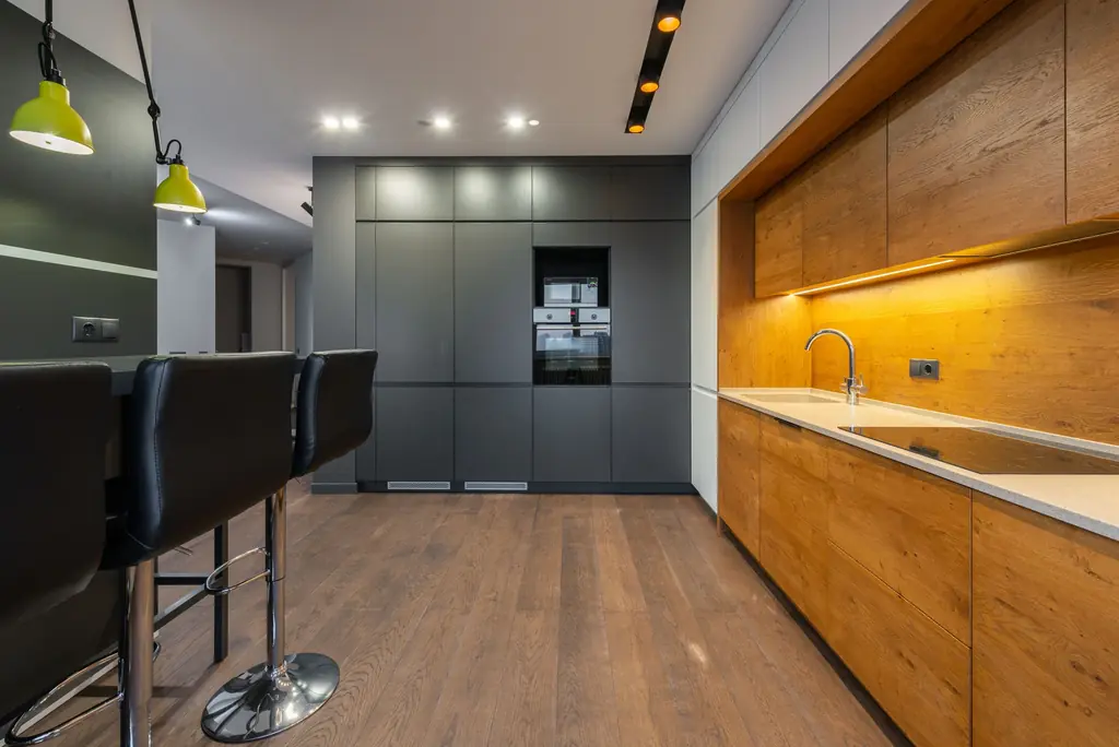 A long galley kitchen with warm oak cabinetry lit from beneath, matte charcoal tall units and a pale oak floor.