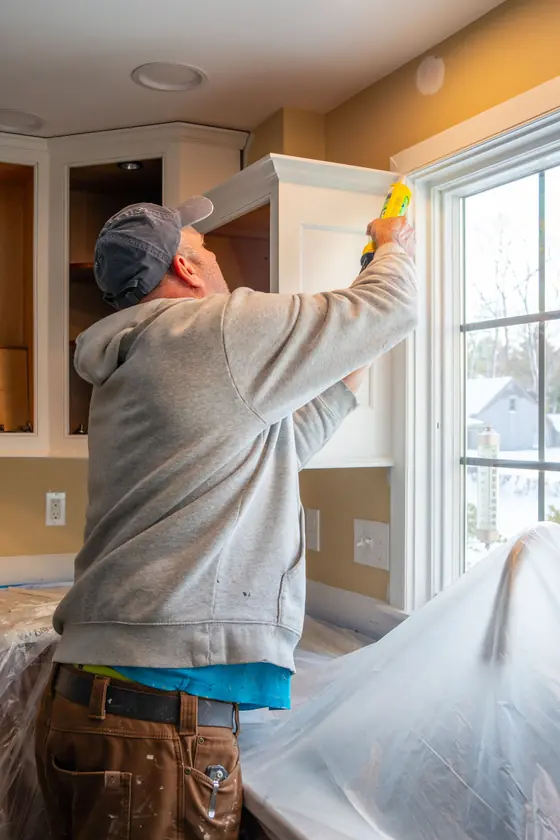 A carpenter running a bead of caulk along a newly installed white upper cabinet beside a window.